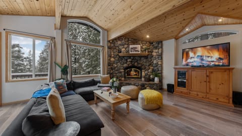 Cozy living room with vaulted wood ceilings, a stone fireplace, and forest-view windows.