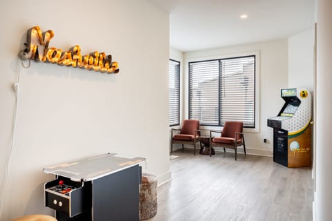 An entertainment room with an arcade machine and a lit "Nashville" sign on the wall, and two burgundy chairs by a large window.