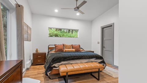 Primary bedroom with a dark upholstered bed, light wood bench, and forest view.