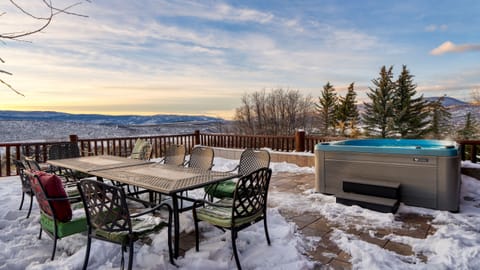 Outdoor dining table and hot tub on a snowy wooden deck with a scenic view at sunset.