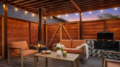 A cozy, covered outdoor lounge featuring wooden paneling, string lights, a BBQ grill, and an outdoor fireplace for evening gatherings.