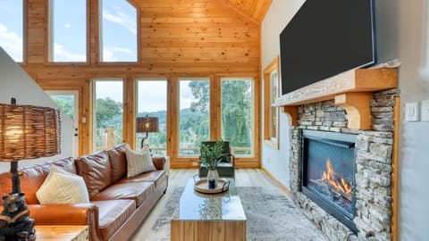 Vaulted living room with floor-to-ceiling windows, a stone fireplace, and warm wood finishes—perfect for cozy nights and scenic mountain views. (Perfect Too)