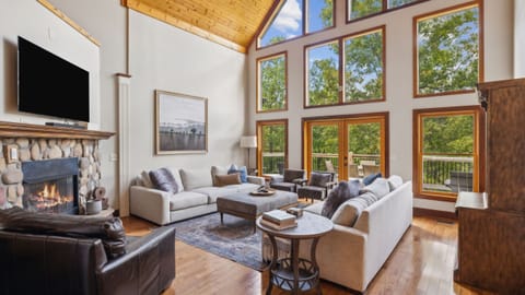 Soaring living room with stone fireplace, wall of windows, and wooded views.