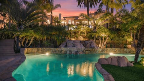 Resort‑style backyard with a glowing pool, lush palms, and a grand estate backdrop.
