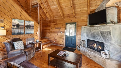 Warm cabin living room with a glowing stone fireplace, a queen sleeper sofa, and sunlight pouring through big mountain‑view windows. (Maggie's Cabin)