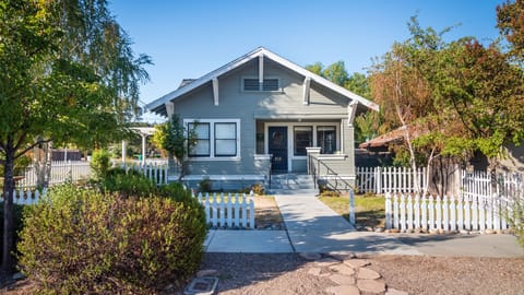 A charming gray cottage welcomes guests with a white picket fence, sunny garden, and inviting front walkway.