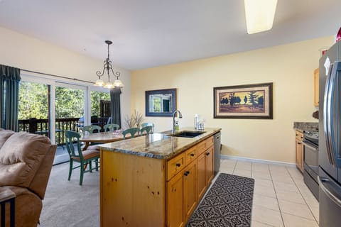 A kitchen with a granite island, wooden cabinetry, stainless steel appliances, and a living area in the background. A dining table with green chairs is near a large window opening to a balcony.