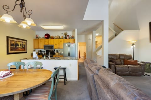A living space with an open layout featuring a kitchen with wooden cabinets, a dining table with chairs, and a cozy seating area. Stairs lead to the upper floor on the right.