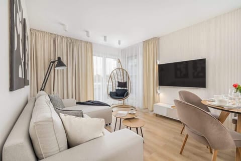 A bright living room with a beige sofa, a TV mounted on the wall, and a hanging chair near the window.
