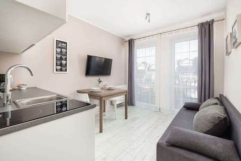 A well-lit living area with a gray sofa, a kitchen counter, and large windows.

