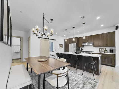 A modern kitchen with dark wood cabinets, white countertops, and an island with bar stools, adjacent to a dining area with a wooden table, white and black chairs, and a green patterned rug.