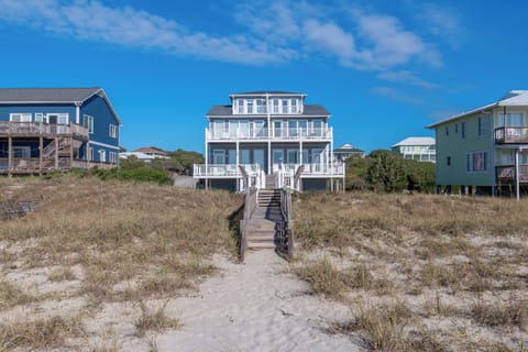 Coastal vacation homes line the dunes with direct beach access via wooden walkway under bright blue skies.