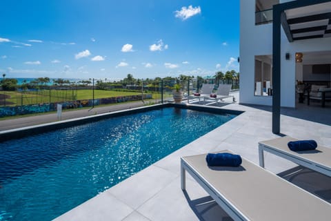 Sparkling pool under a blue sky, adjacent to a contemporary house, with lounge chairs, offering a stunning view of the ocean and an artistic wall.