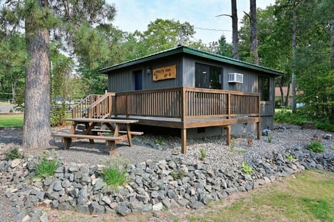 A small, grey wood-sided cabin with a large wooden deck and picnic table, situated on a rocky landscape with tall pine trees.
