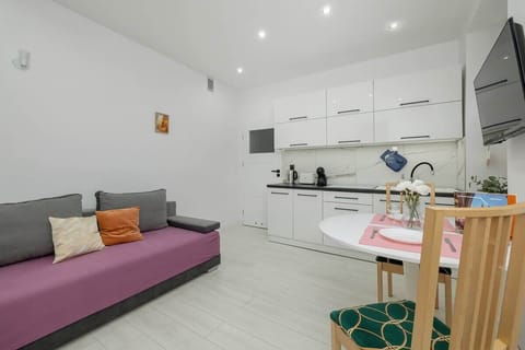 Living room with a modern, minimalistic design. White walls, recessed lighting, and a pink sofa with green plant decor in the corner. The room appears bright and open, with a simple, clean aesthetic.