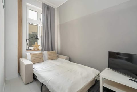 A bright bedroom with a single bed, a small bedside table, and a window draped in light curtains.
