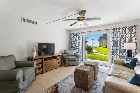 - Cozy living room with plush seating, a ceiling fan, & a flat screen TV for relaxing evenings
- Sliding doors reveal beautiful green lawns & glimpses of palm trees just outside
- Natural light brightens the space for a breezy coastal vibe