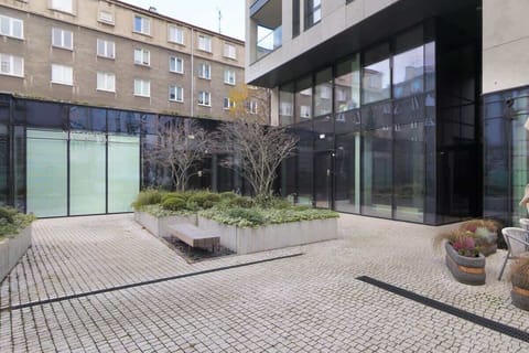 An outdoor courtyard with a paved walkway and open seating area, framed by contemporary building facades.

