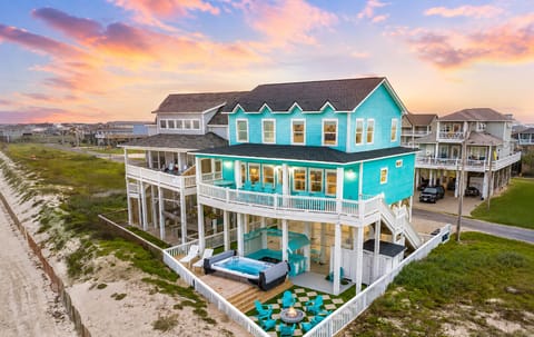 Aerial view of a turquoise beachfront home with Gulf-facing decks, a private swim spa, fire pit lounge, fenced outdoor area and direct access to the sandy Bolivar Peninsula beach.