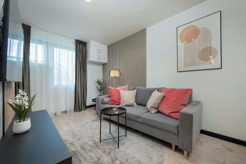 A modern, stylish living room featuring a gray sofa with pink cushions, a round coffee table, and minimalist decor. A large window brings in ample natural light.