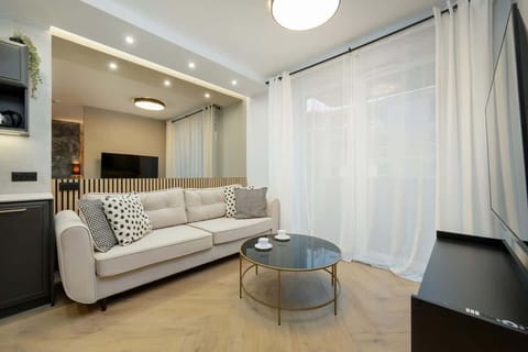 A modern living room with a light beige sofa, round glass coffee table, and gold accents, framed by sheer white curtains for a bright, airy feel.


