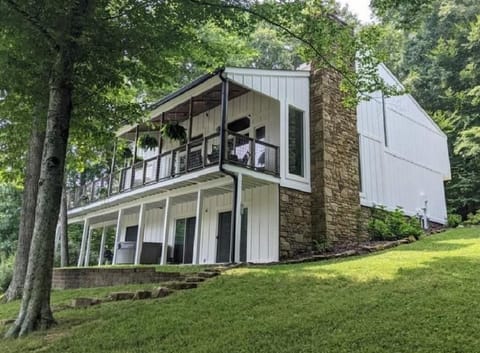 Two-story exterior with white vertical siding, stone chimney, and covered upper balcony overlooking the yard.