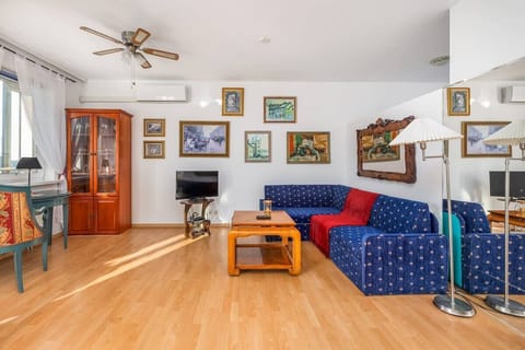 A bright living room with a wooden floor, a blue patterned sofa, multiple framed artworks on the walls, and a ceiling fan providing a cozy atmosphere.


