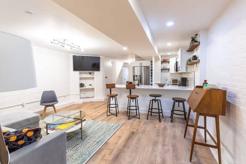 Living room with a queen size sofa bed, coffee table and additional seating. A work desk is available for remote work. Bar stools sit before the kitchen and a wall mounted TV sits above the built in storage. 