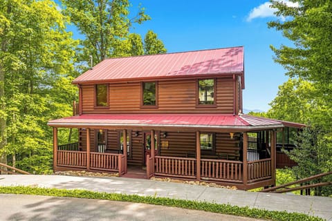 A charming two-story log cabin with a wrap-around porch nestled among towering trees, offering a peaceful mountain retreat with ample parking and natural surroundings.