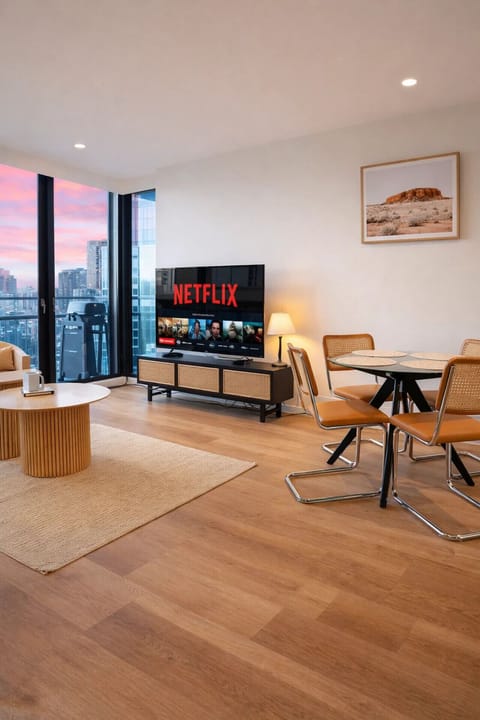 An open living and dining space with warm timber floors, rattan furnishings, and a large smart TV facing floor-to-ceiling windows. A vivid city sunset fills the room with colour, creating a relaxed evening atmosphere.