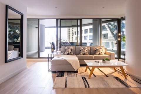 Generous living room with an L-shaped sofa, timber coffee table, and floor-to-ceiling glass doors opening to a wide balcony. A large black-framed mirror and warm timber floors add depth and light to the space.  *(209 characters)*