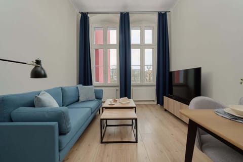 A cozy living room with blue sofas, a wooden coffee table, and a flat-screen TV placed against the wall. The large windows with dark blue curtains allow ample natural light to fill the room.
