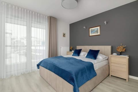 A modern bedroom with gray accent walls, a neatly made double bed with blue bedding and pillows, and a bedside table with a decorative plant and framed artwork on the wall.

