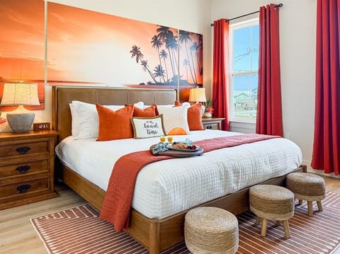 Bright coastal bedroom with a king bed, sunset palm artwork, warm coral accents, bedside lighting, and large windows in a Galveston vacation rental.