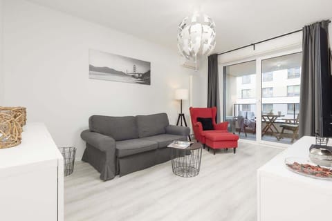 A bright living room with a grey sectional sofa, a bold red armchair, a glass coffee table, and large windows overlooking the balcony, creating a modern and inviting space.
