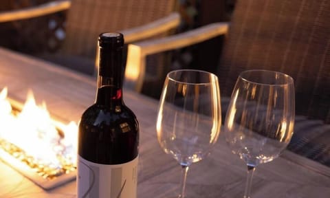 This outdoor space is a perfect evening setting, featuring a rustic wooden table with an integrated fire pit, accompanied by a bottle of wine and glasses.