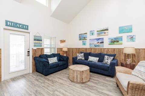 Sink into plush navy sofas surrounded by cheerful beach artwork in this bright, coastal-themed living space with bamboo accents and vaulted ceilings.