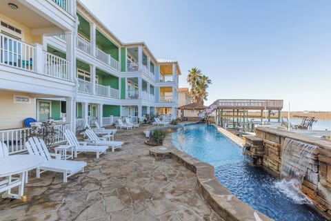 Colorful waterfront resort featuring multiple buildings with balconies, swimming pool with waterfall, and lakefront access under clear skies.