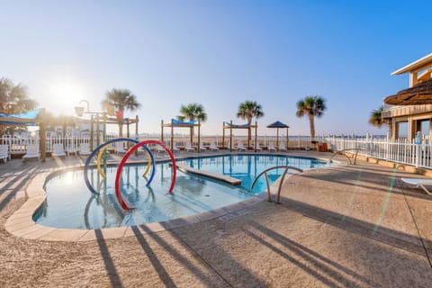 Beachfront resort pool area with palm trees and children's play features under golden morning sunlight.