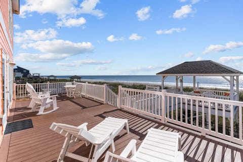 Large Oak Island, NC beachfront deck with white lounge chairs, rockers, dining seats, gazebo, ocean views, beach dunes, railings, and sunny coastal sky.