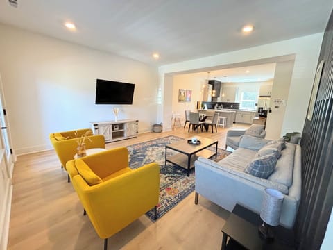 LIVING ROOM – This spacious and modern open-concept living area is designed for both comfort and style. The bold yellow armchairs provide a striking contrast to the sleek black accent wall, while the soft gray sectional offers ample seating for relaxation. The room flows seamlessly into the dining and kitchen areas, making it perfect for entertaining. The colorful area rug adds...