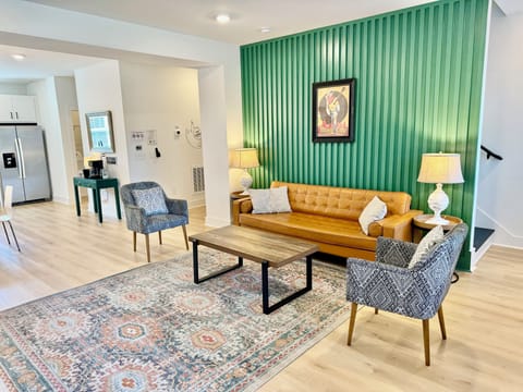 LIVING ROOM – This vibrant living room is styled with a mix of modern and vintage elements, creating a cozy yet contemporary atmosphere. The bold green accent wall features iconic local artwork, adding a touch of Nashville's character to the space. A mustard-yellow leather sofa and patterned chairs invite you to relax in style, while the nearby kitchen and dining area provide a...