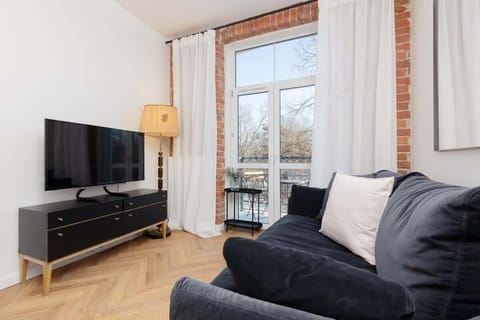 A modern living room with a navy blue sofa, white cushions, and a minimalist TV stand, complemented by a brick accent wall and large windows with sheer curtains.
