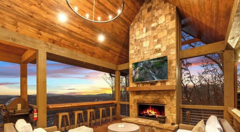 Stunning mountain views frame this covered outdoor living space with stone fireplace, cozy seating, and bar stools—perfect for relaxing evenings under soaring wood ceilings as the sun sets beyond the hills.