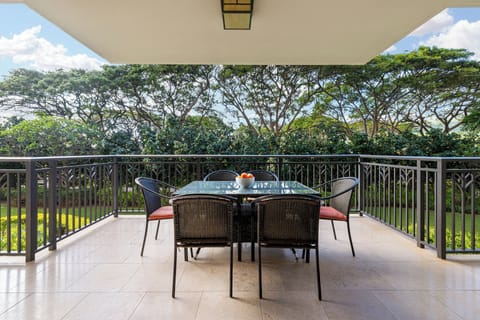 A covered balcony featuring a glass-top dining table set for four, offering peaceful views of lush tropical trees and manicured grounds.