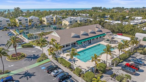 Aerial view of a community center with a large outdoor pool, surrounding lounge chairs, several tennis courts, and a parking lot. Residential buildings and lush greenery are visible in the background.