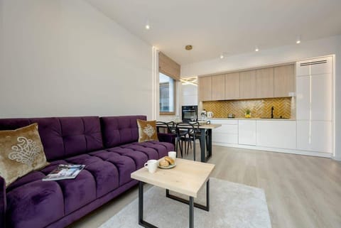 A spacious living room featuring a large purple sectional sofa, a modern coffee table, and a mounted TV. The open-plan layout seamlessly connects the living area with the kitchen, which is equipped with light wood cabinetry and contemporary fixtures.