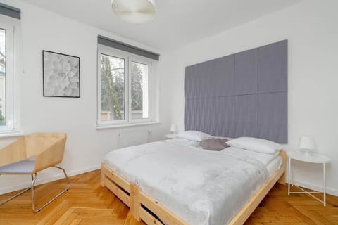 A bright bedroom with a wooden bed frame, a soft gray headboard, and large windows letting in natural light.
