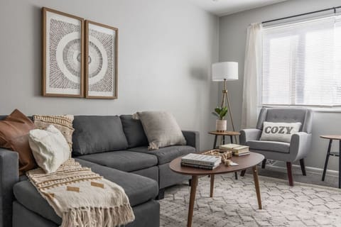 Stylish living room with a plush sectional, cozy accent chair, and soft layered textures. Bright natural light, modern decor, and a warm neutral palette create the perfect space to relax, read, or unwind after a busy day.