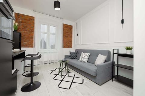 A modern open-plan living space with a gray sofa, a black bar-style dining table, and white walls with exposed brick accents, creating an industrial-chic ambiance.
